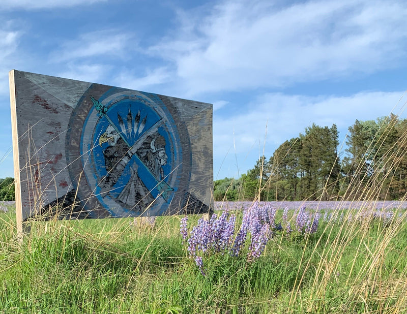 Prairie Island Reservation's symbol on a billboard in a grassy field with purple Lupine flowers, trees, and blue sky in the background